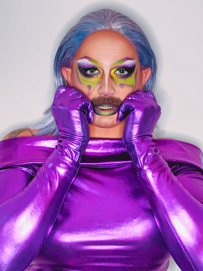 Bust view of Halal Bae wearing a shiny purple catsuit, looking into the camera with their hands curled up by their cheeks.