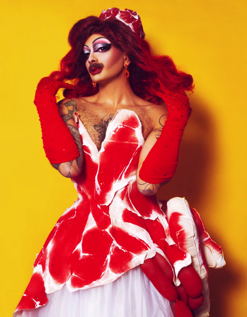 Halal Bae posing like a diva tossing her hair back while wearing a meat-themed dress and red rhinestone opera gloves.