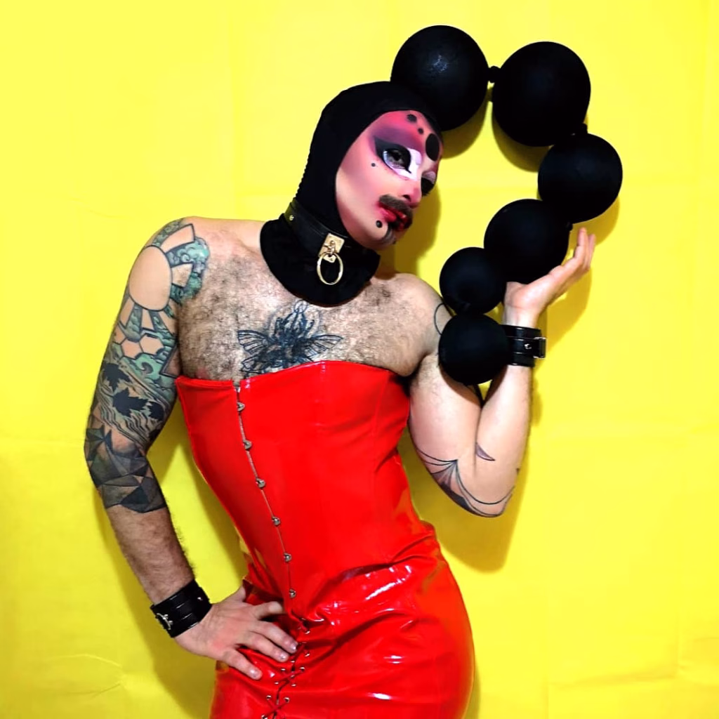 Halal Bae posing dramatically in a shiny red leather corset dress and a black headpiece that looks like a string of large beads.